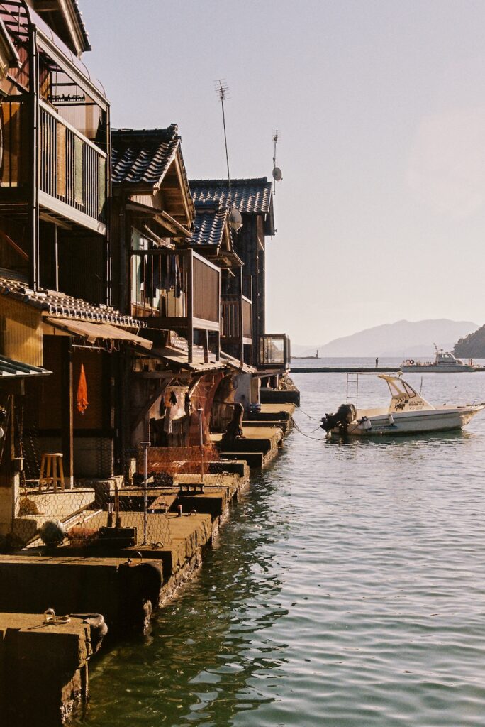 Traditional wooden houses built directly along the water in a Kyoto fishing town, with boats floating nearby and soft light reflecting off the calm surface.