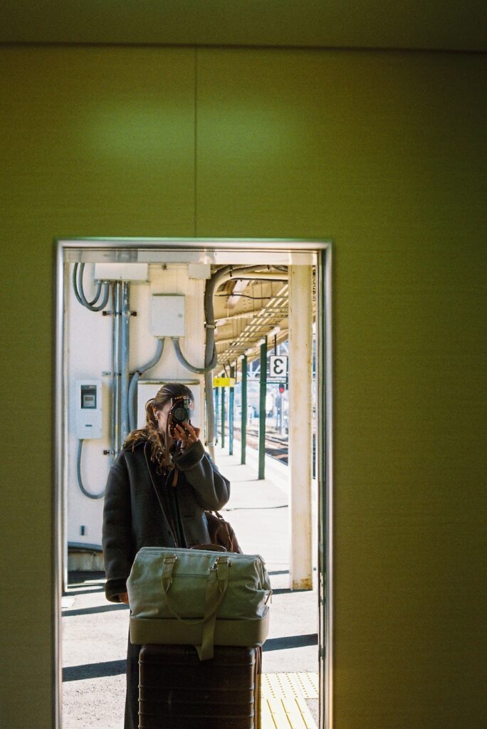 A traveler reflected in a station mirror in Kyoto, standing beside luggage on the platform with trains and overhead lines visible beyond the frame.