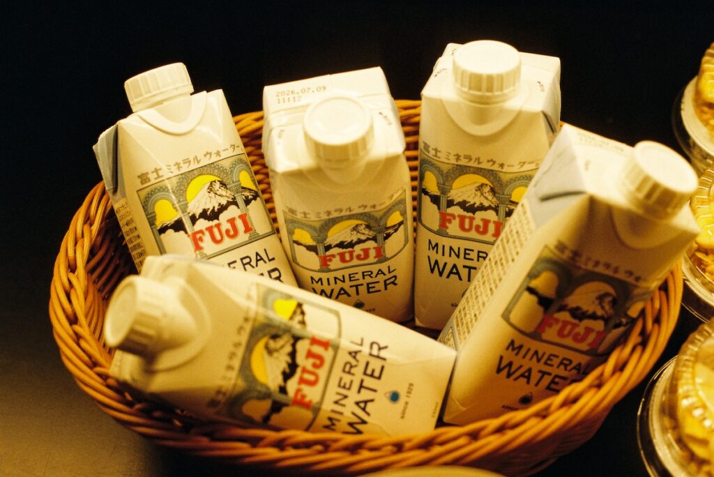 Cartons of Fujisan mineral water arranged in a woven basket, featuring illustrated Mount Fuji labels in warm indoor light.
