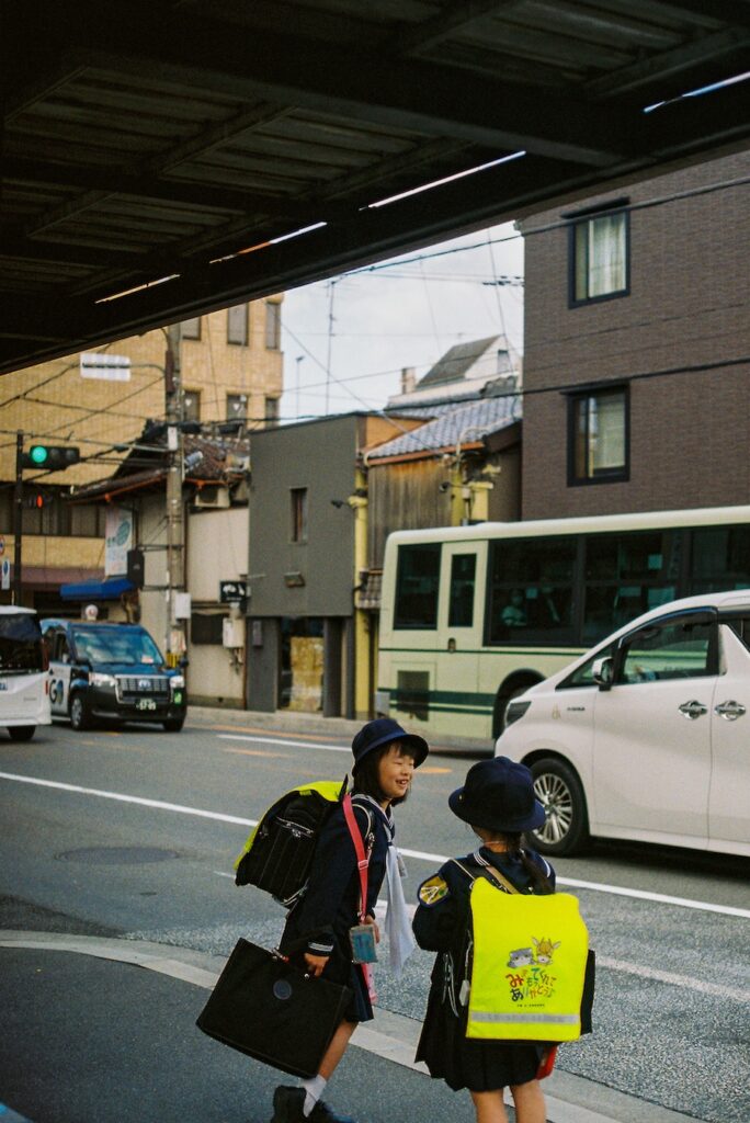Two schoolchildren wearing bright yellow backpacks cross a quiet residential street in Japan, surrounded by compact buildings, parked cars, and soft afternoon light.