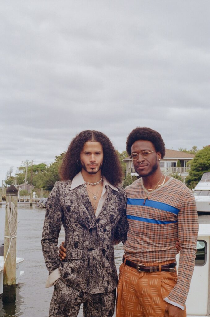 Two friends standing beside the harbor in Fire Island Pines with boats and waterfront homes in the background.